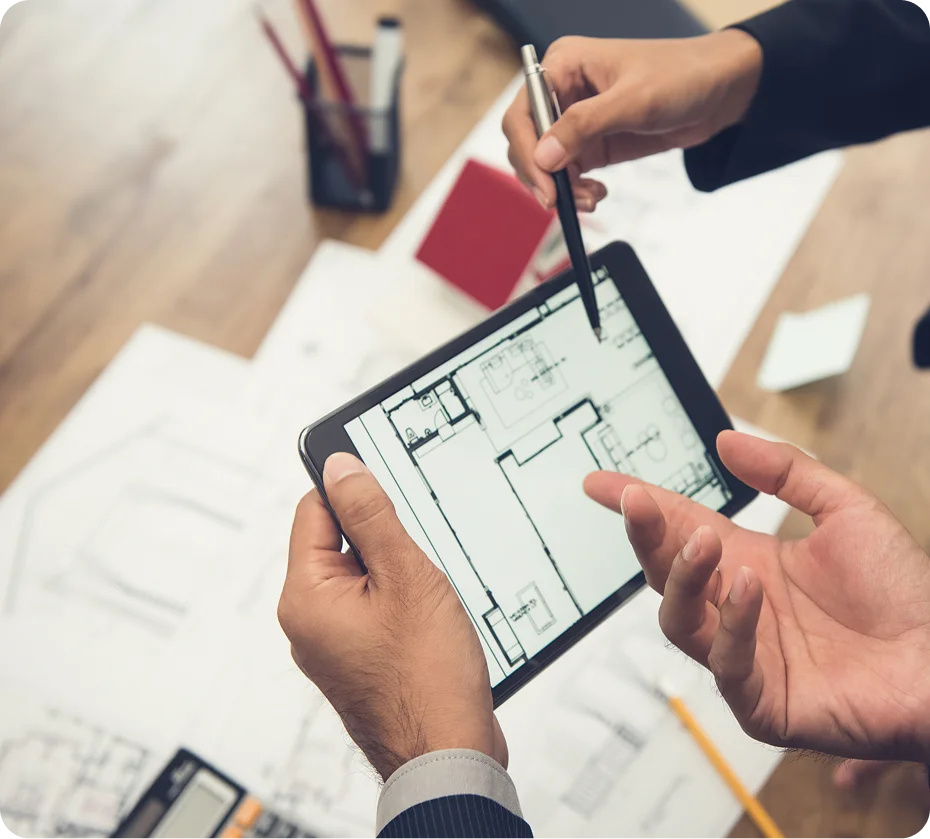 GettyImages-1279995963 (1) Two hands holding a tablet displaying architectural plans. Another hand gestures with a pen, pointing at the screen. Papers are scattered on the table below.