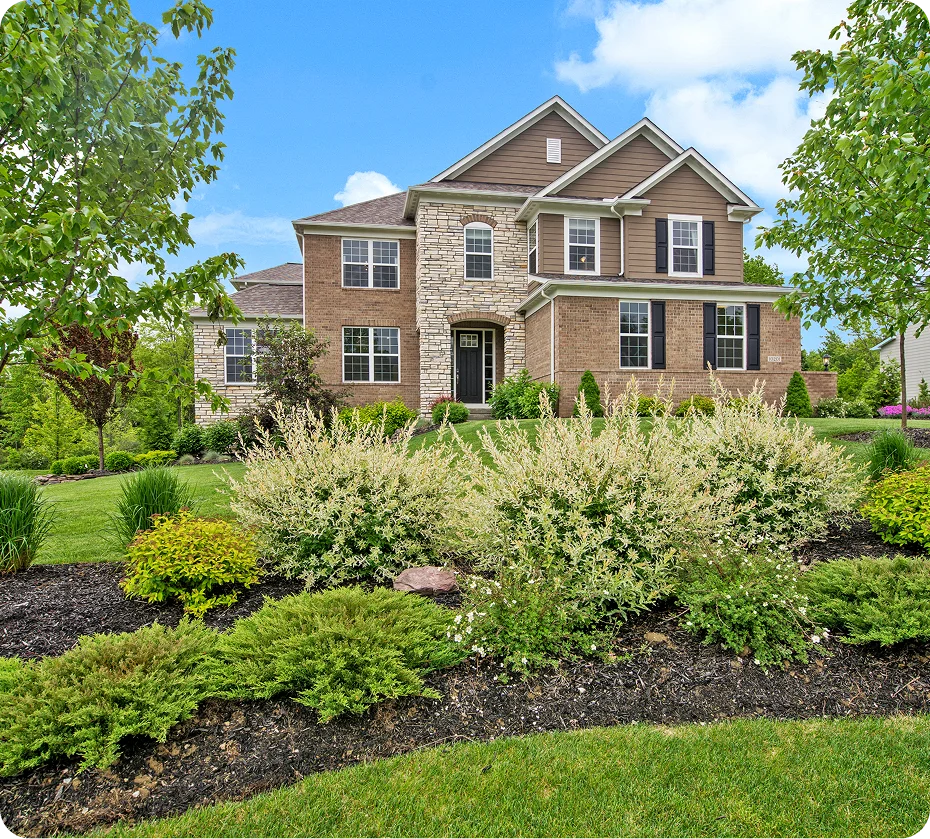 GettyImages-1279995963 (4) A two-story brick and stone house with a manicured lawn and landscaped shrubs, under a clear blue sky.