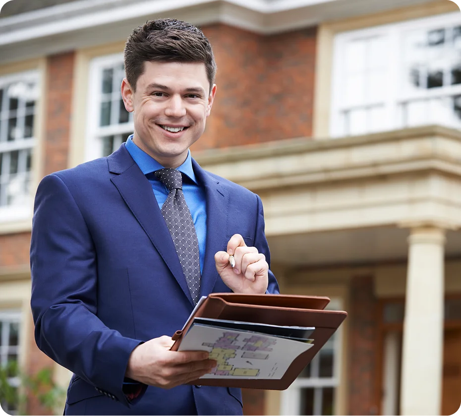 GettyImages-1279995963 A man in a blue suit smiles while holding documents and a tablet, standing in front of a brick house.