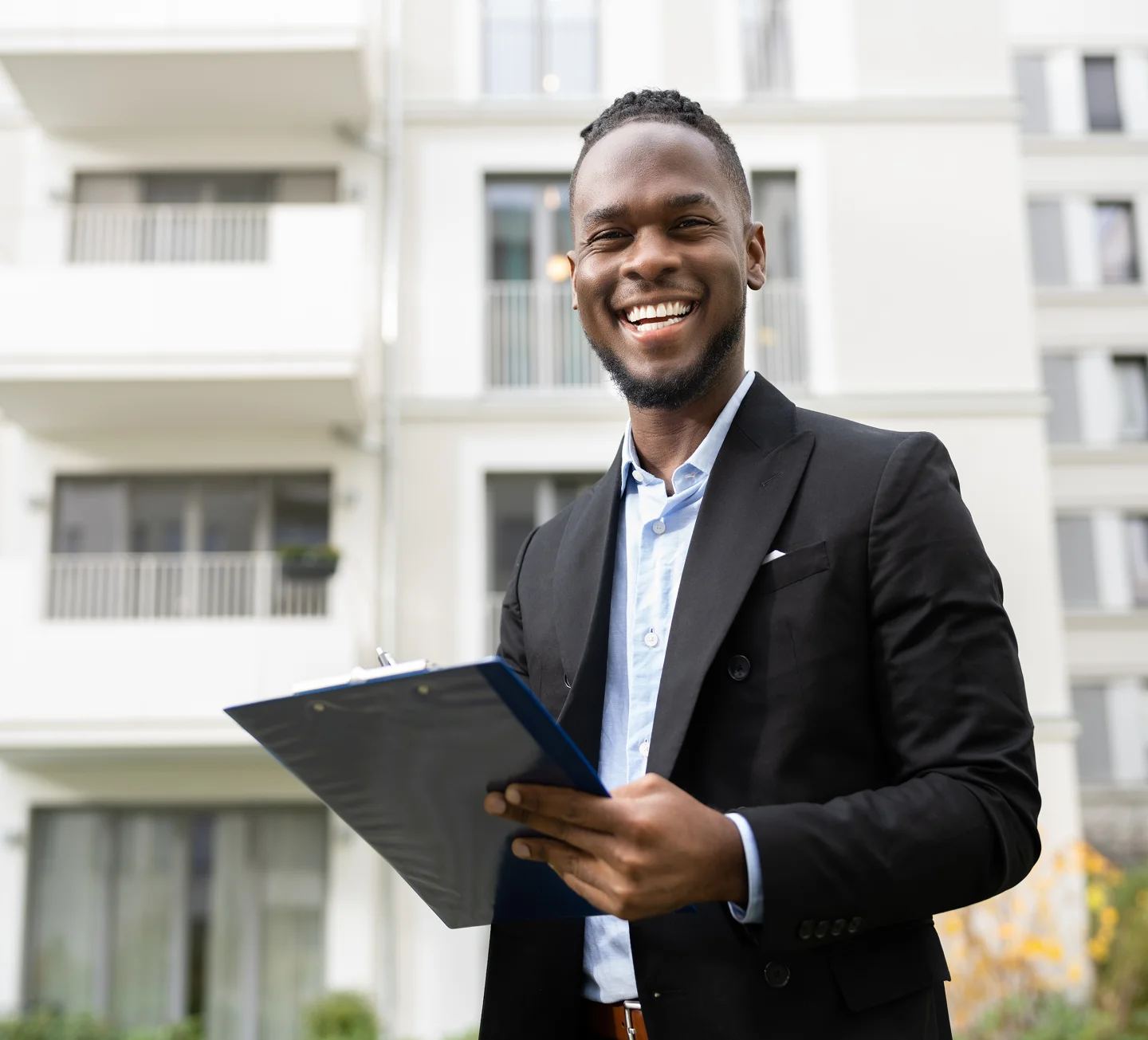 GettyImages-1481196113 (1) Smiling man in a suit holds a clipboard, standing outdoors in front of a modern apartment building.