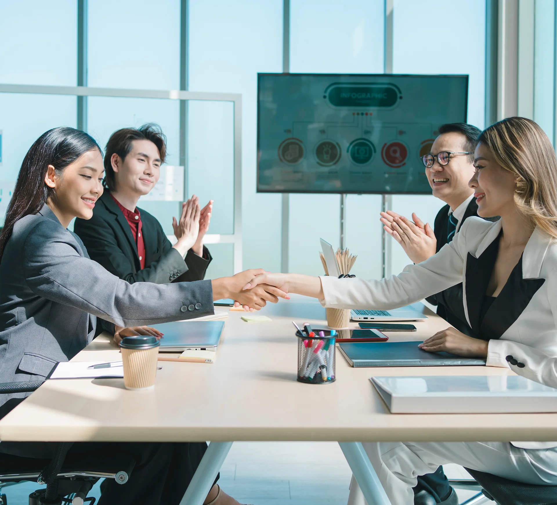 GettyImages-1481196113 Two people shake hands across a meeting table. Two others are seated next to them, clapping. Laptops and cups are on the table. A presentation screen is in the background.